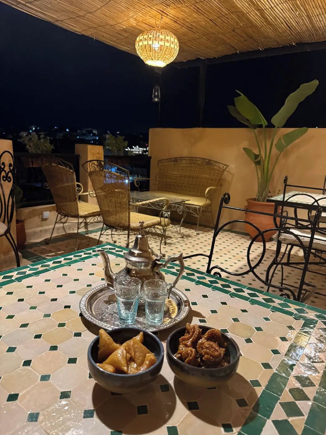An outdoor dining area with a table set for two, featuring a traditional metal tea set, two glasses, and two bowls of food.