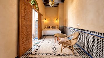 Un pasillo estrecho que conduce a un dormitorio con una cama, dos luces fijadas a la pared, una alfombra estampada y una silla de mimbre.