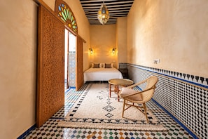 A narrow hallway leading to a bedroom with a bed, two wall-mounted lights, a patterned rug, and a wicker chair. - Riad La Petite Maison (Fès)