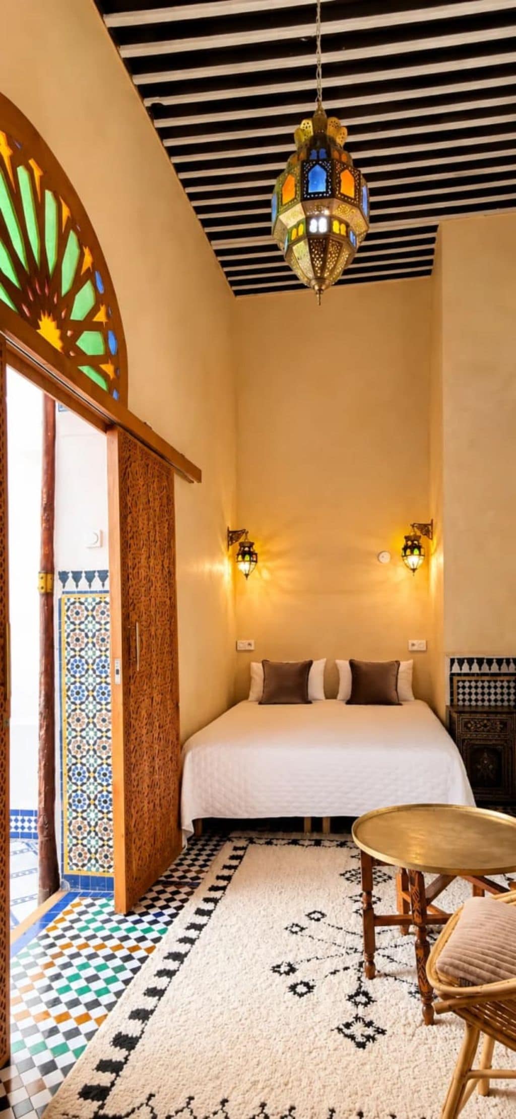 A room with a bed, two wall-mounted lights, a hanging lantern, a patterned rug, and a small round table.
