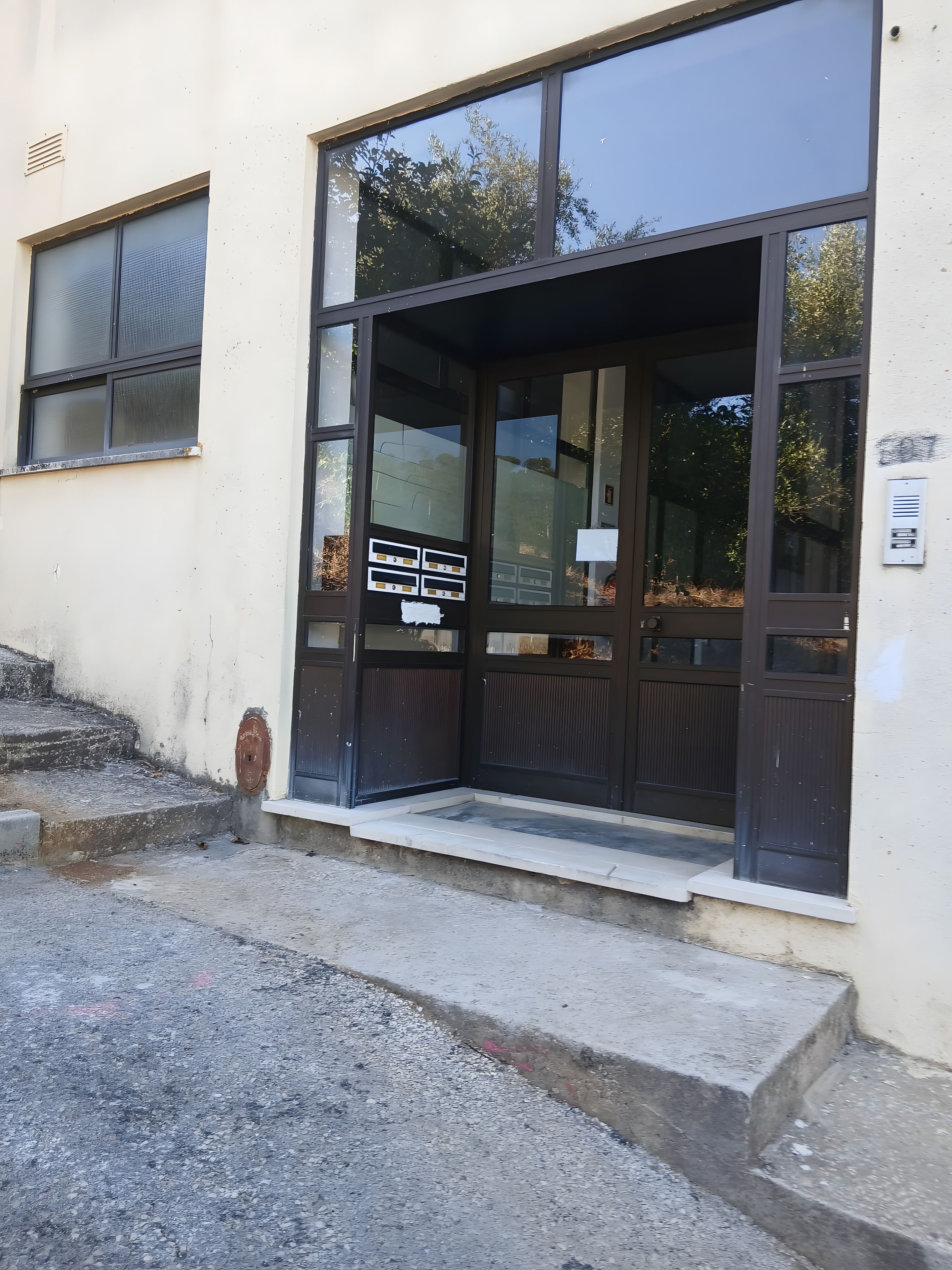L’entrée d’un immeuble se compose d’une grande porte vitrée, d’une échelle de secours en métal et d’un petit espace extérieur avec des marches.