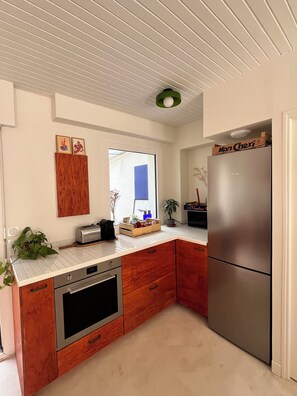 A compact kitchen with wooden cabinets, a stainless steel refrigerator, and a window with a view. - Three-bedroom house with a garden overlooking the forest. Quiet neighborhood, near a bike path (Capbreton)