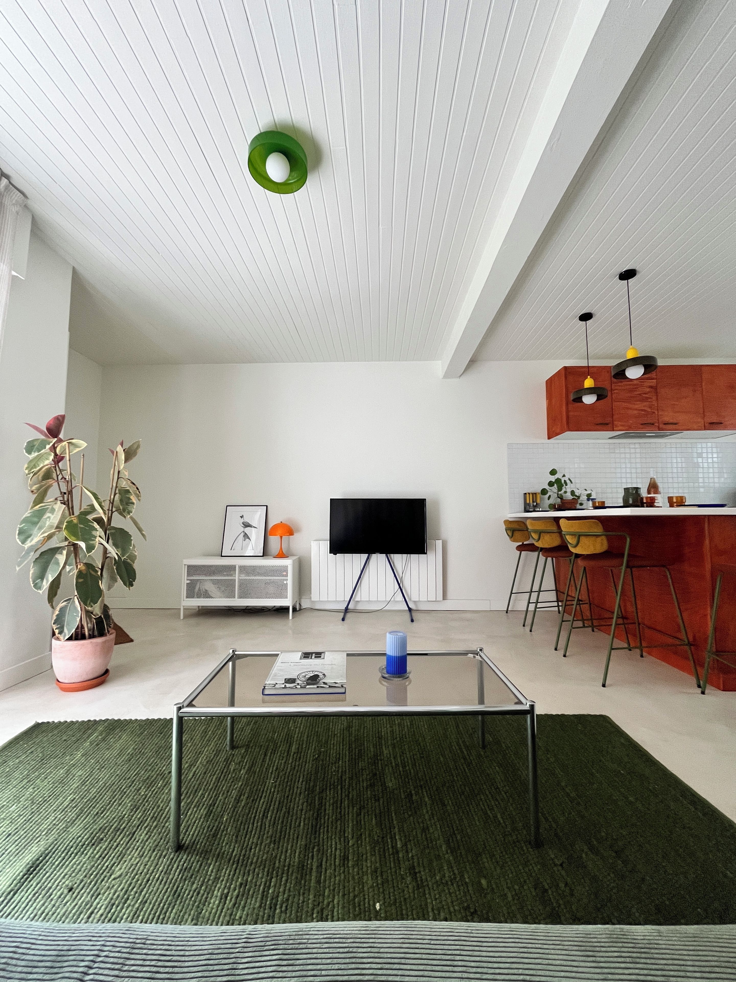 Una sala moderna con una mesa de centro de vidrio, una planta en maceta, un televisor de pantalla plana y una cocina con armarios de madera y taburetes de bar.