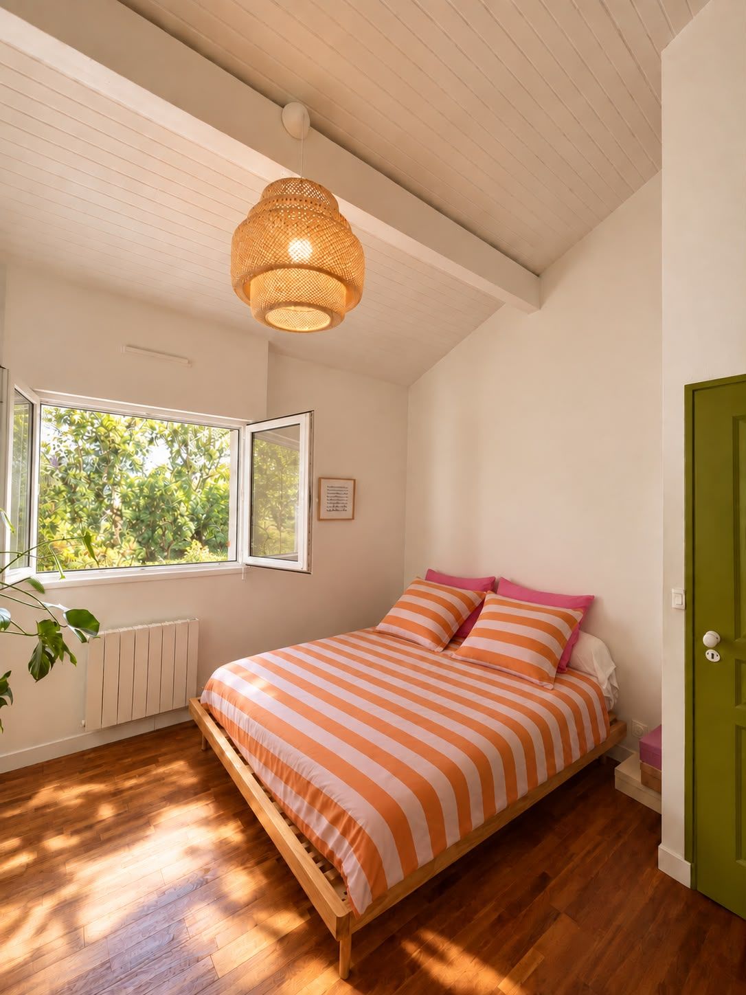 A bedroom with a wooden bed, a window with a view of trees, a green door, and a woven light fixture.