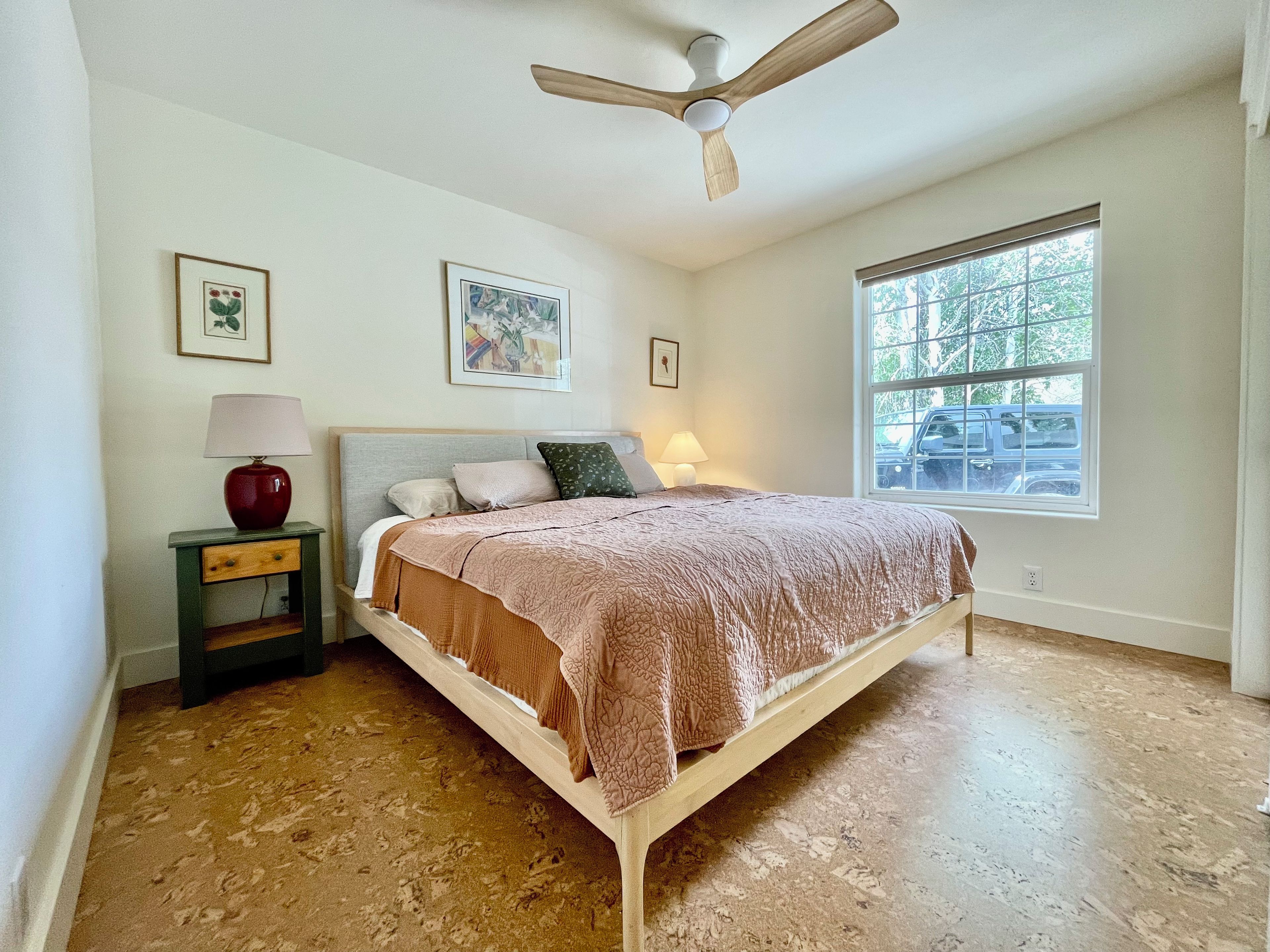 Un dormitorio con cama, mesita de noche, lámpara, ventilador de techo y ventana con vistas al exterior.