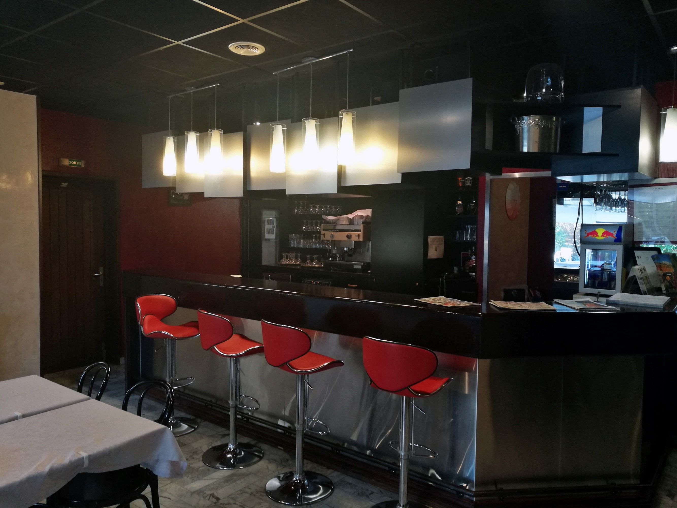 Un espace bar avec un comptoir, des tabourets de bar rouges, des suspensions décoratives et un réfrigérateur orné du logo Red Bull.