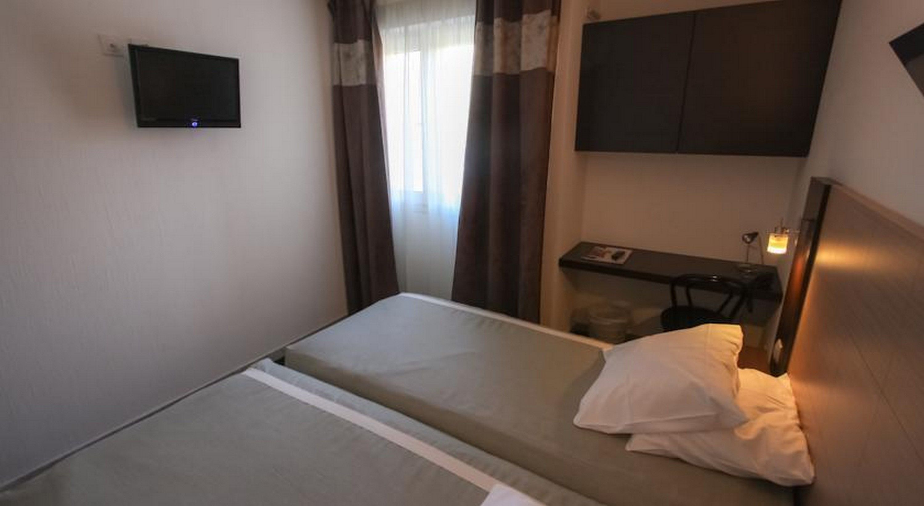 Une petite chambre d’hôtel simple, avec un lit, un bureau, une chaise, une télévision et une fenêtre avec des rideaux.