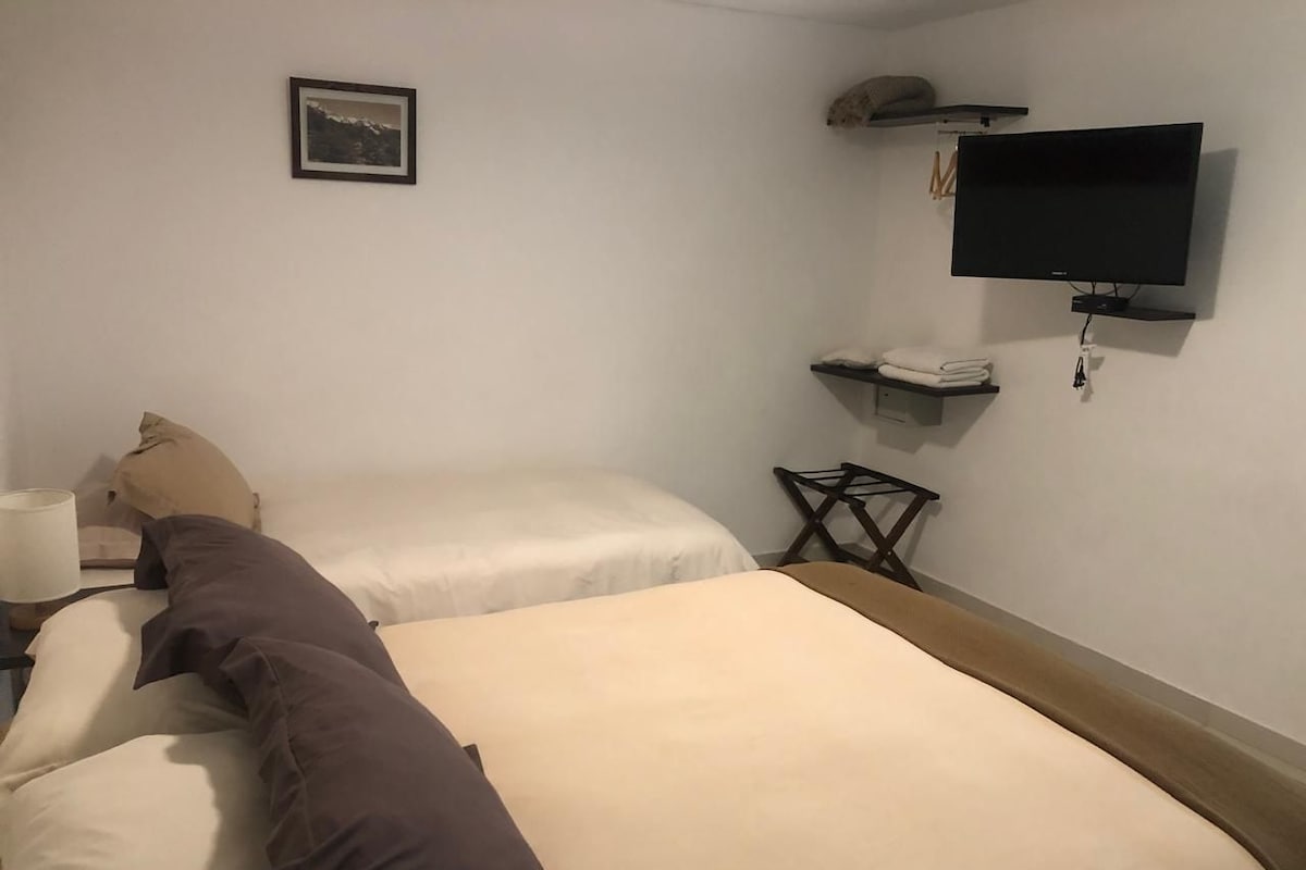 A hotel room with two beds, a flat-screen TV mounted on the wall, a small table with a lamp, and a framed picture on the wall.