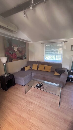 A living room with a sectional sofa, a glass coffee table, a painting on the wall, and a window with blinds. - Stunning, fully equipped mobile home in a very quiet community (Sainte-Maxime)