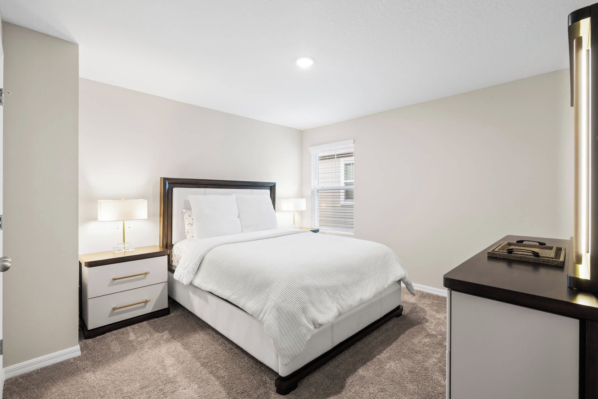 A bedroom with a bed, bedside tables, a nightstand, a window with blinds, and a mirror.