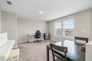 Una habitación pequeña y bien iluminada con kitchenette, una silla de oficina, una mesa de comedor con sillas y un ventanal con persianas.