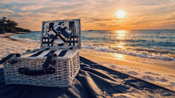 A wicker picnic basket with a black handle on a beach with the ocean and a sunset in the background. - Charming Home Near Macdill AFB Hot Tub Fire Pit (Tampa)