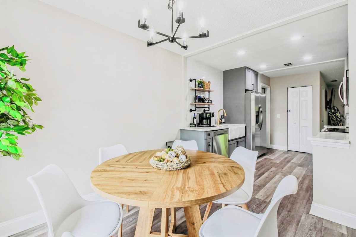 A dining area with a round wooden table surrounded by white chairs, a chandelier, and a kitchen in the background.