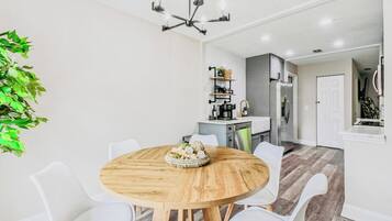 A dining area with a round wooden table surrounded by white chairs, a chandelier, and a kitchen in the background.