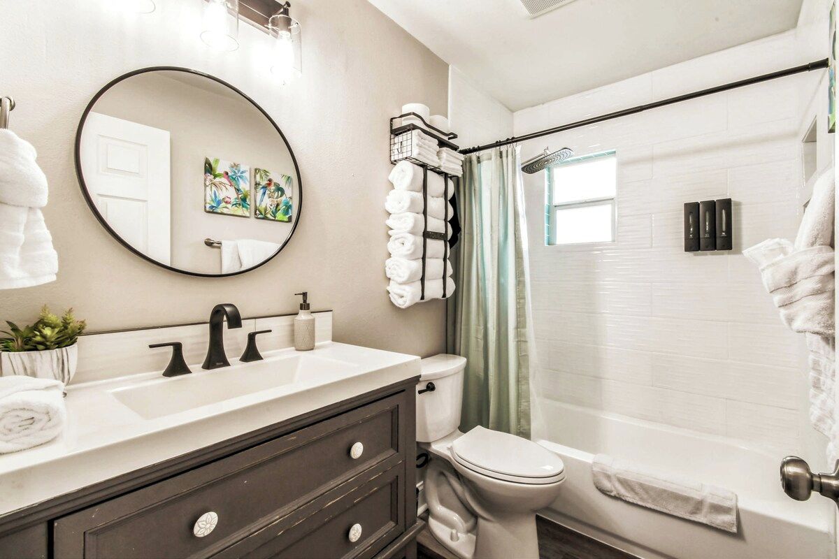A bathroom with a round mirror, a toilet, a sink with a dark cabinet, and a shower area with a curtain.