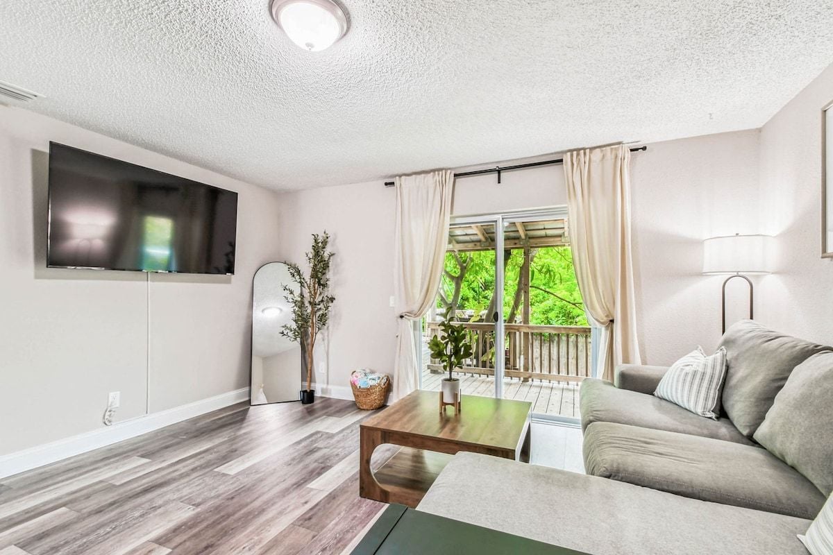 Ein Wohnzimmer mit Sofa, Couchtisch, Wand-Fernseher, einem Fenster mit Vorhängen und einem Balkon mit Geländer.