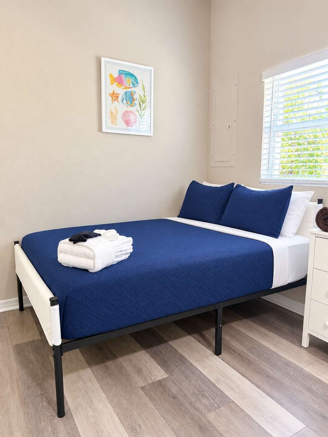 A bedroom with a bed, a window with blinds, a wall-mounted artwork, and a bedside table.