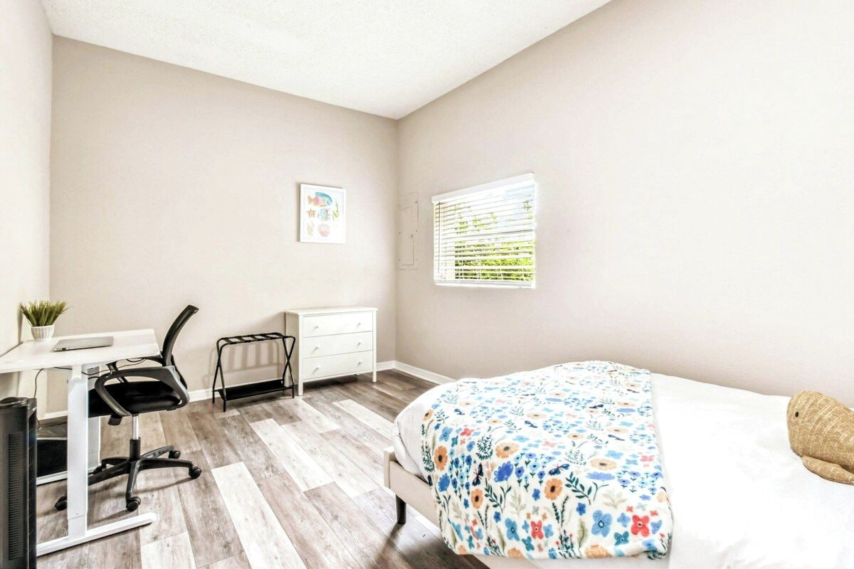 A bedroom with a bed, a desk, a chair, a small table with a plant, and a window with blinds.