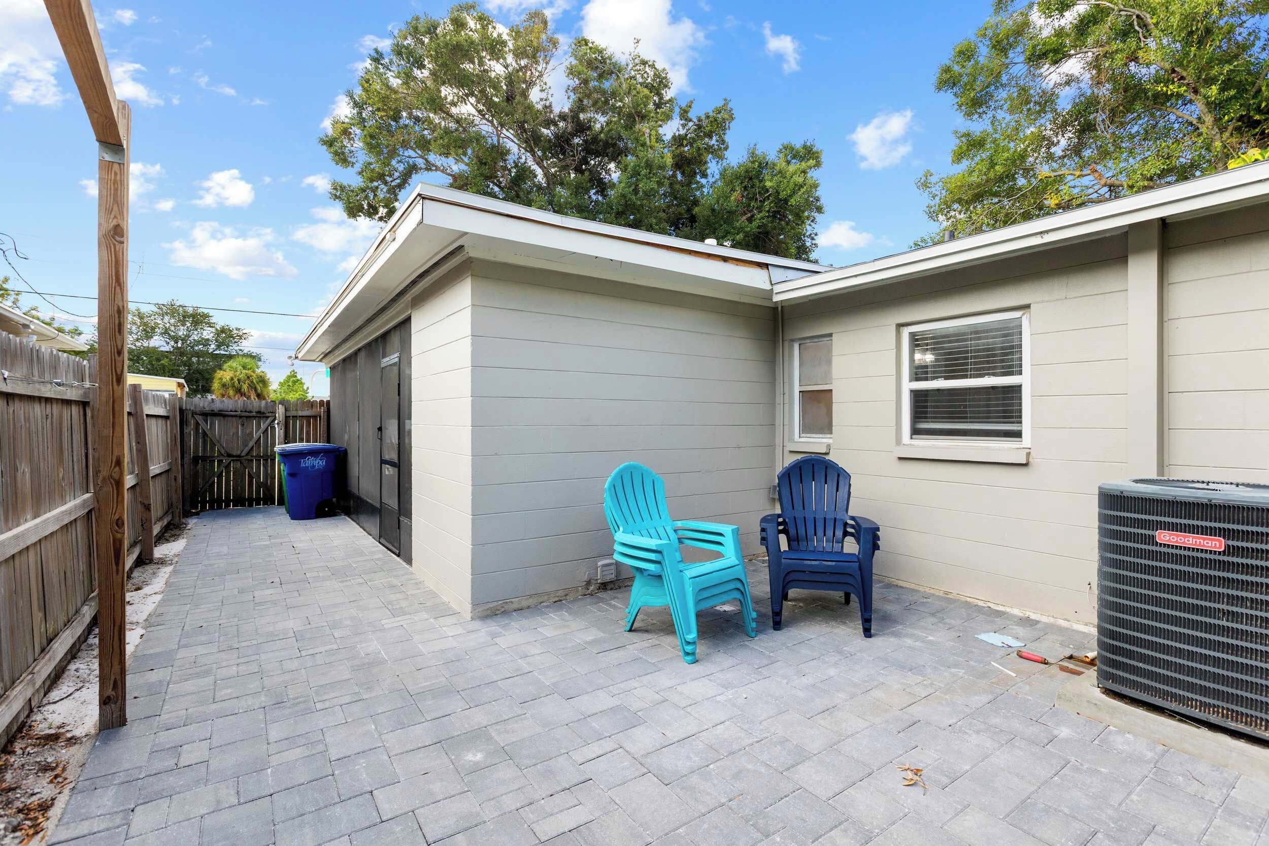 A residential backyard with a patio, two Adirondack chairs, a trash bin, and a large air conditioning unit.