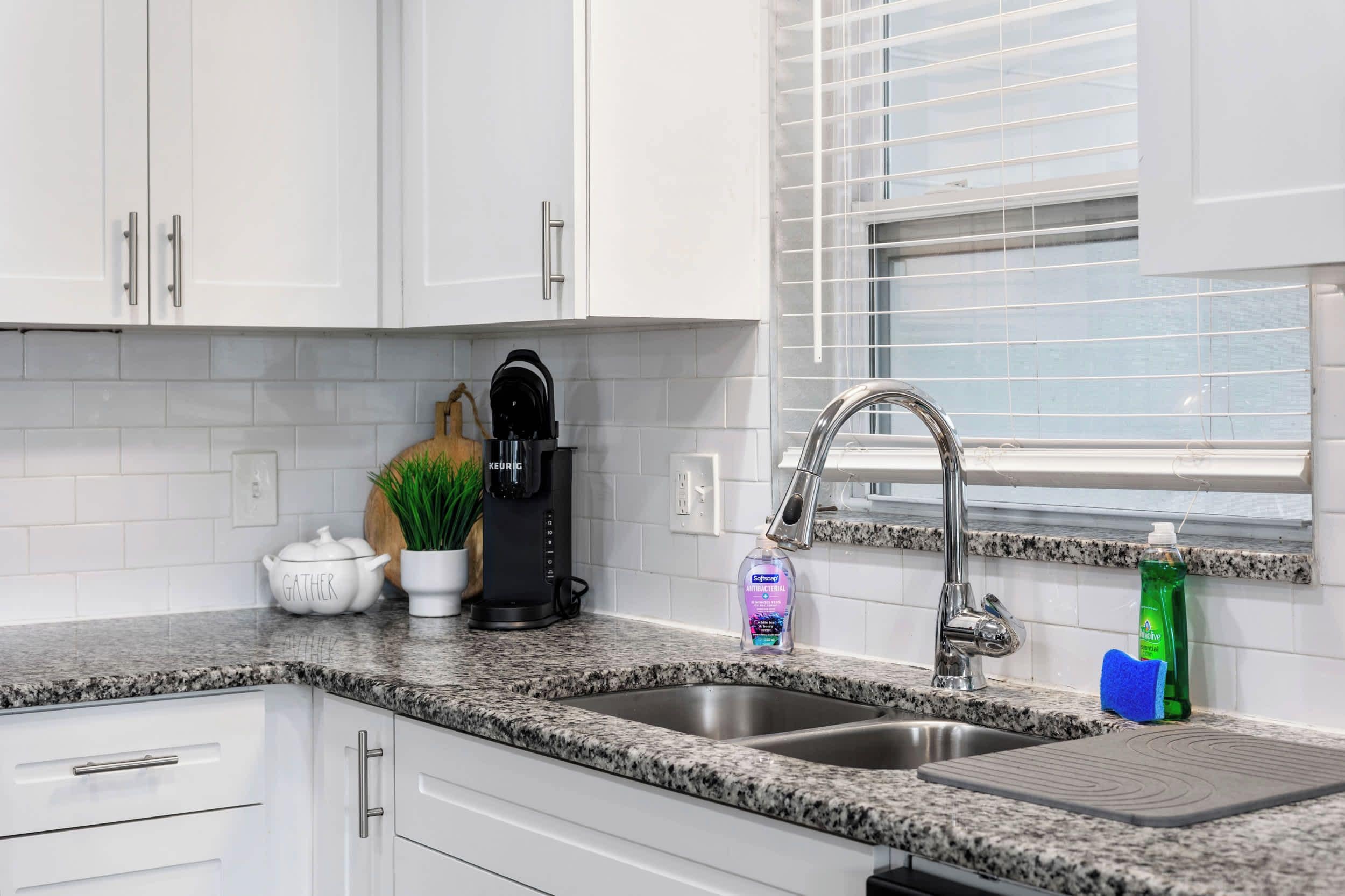A kitchen with white cabinets, a stainless steel sink, a coffee maker, and a window with blinds.