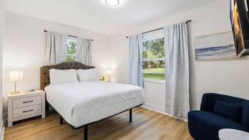 A bedroom with a bed, bedside tables, a nightstand, a sofa, a window with curtains, and a painting on the wall.