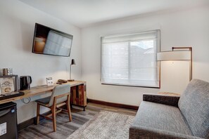 A compact living space with a wooden desk, a chair, a sofa, a television mounted on the wall, and a window with blinds. - Ski-in/Ski-out Breck King Room | Cozy Lounge + Spa (Breckenridge)