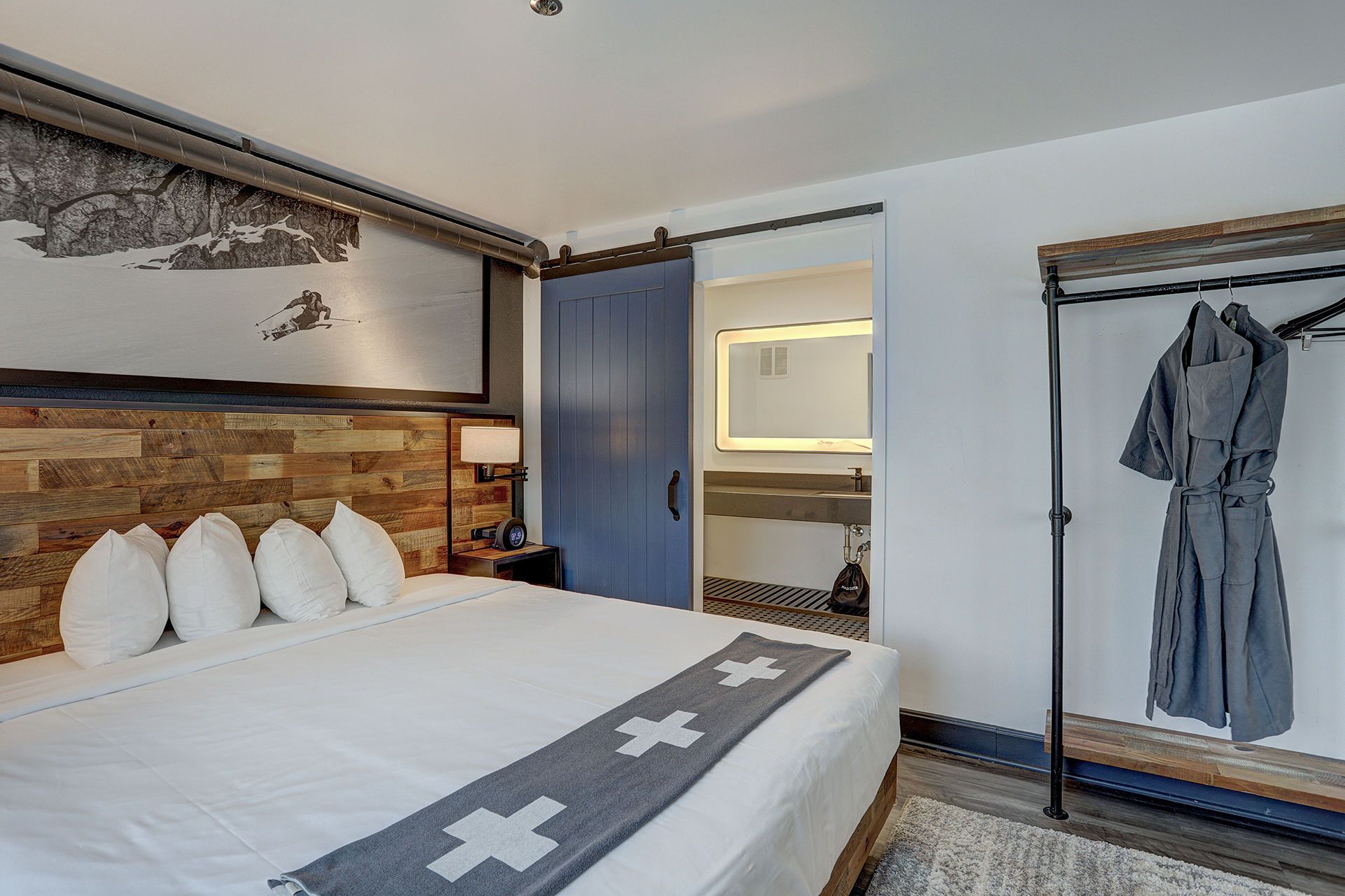Quarto de hotel com cama, cabeceira de madeira, porta-robes e banheiro visível através de uma porta aberta.