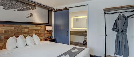 Une chambre d’hôtel avec un lit, une tête de lit en bois, un porte-peignoir et une salle de bain visible à travers une porte ouverte.