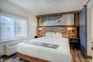 A modern hotel room with a large bed, wooden headboard, two bedside tables with lamps, a large window, and a wall-mounted artwork. - Ski-in/Ski-out Breck King Room | Cozy Lounge + Spa (Breckenridge)