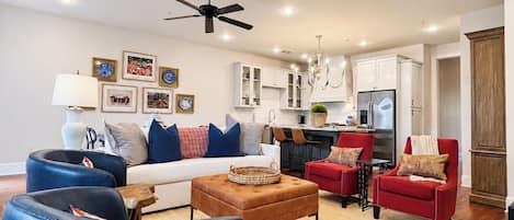 Ein modernes Wohnzimmer mit einer Couch, einem Hocker und Sesseln, mit einer Küche im Hintergrund und einem Deckenventilator.