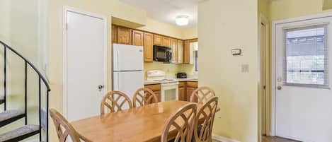 A dining area with a wooden table and chairs, a kitchen with wooden cabinets, a white refrigerator, and a microwave.