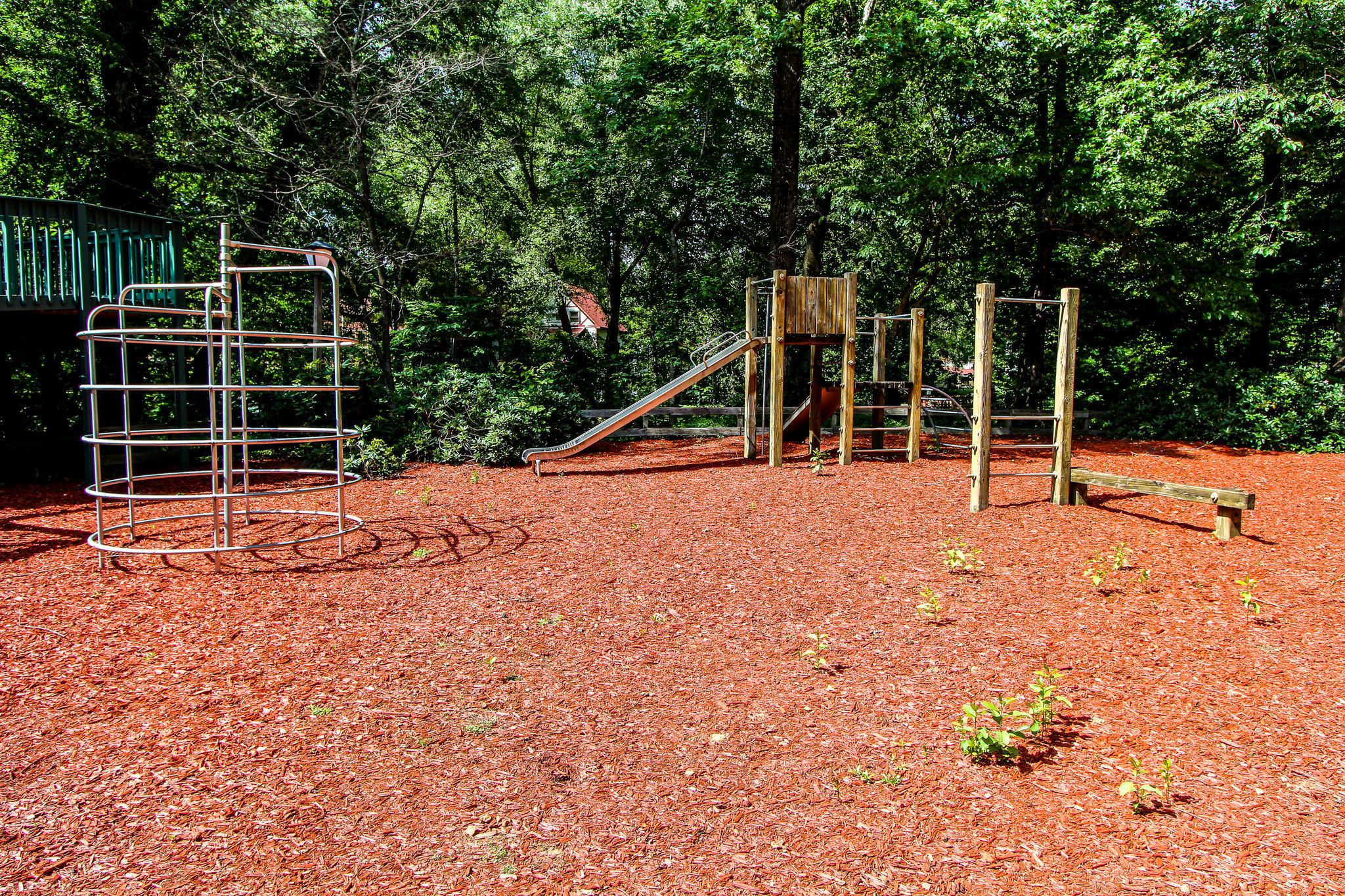 Une aire de jeux avec un toboggan en spirale en métal, une structure d’escalade en bois et un petit toboggan, le tout entouré d’une zone boisée.
