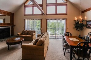 A spacious living room with a fireplace, a sofa set, a dining table with chairs, and large windows offering a view of greenery. - 1BR Condo w/ Private Balcony | Near Alpine Trails! (Helen)
