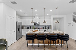 Cozinha moderna com armários brancos, uma ilha central com banquetas, eletrodomésticos de aço inoxidável e iluminação pendente.