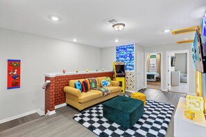 Uma sala de estar com lareira de tijolos, sofá com almofadas coloridas, tapete xadrez e máquina de fliperama Pac-Man.