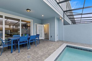 Eine überdachte Terrasse mit Pool, eine Essecke mit blauen Stühlen und einem Tisch sowie eine Glasdoor, die zum Küchenbereich führt.