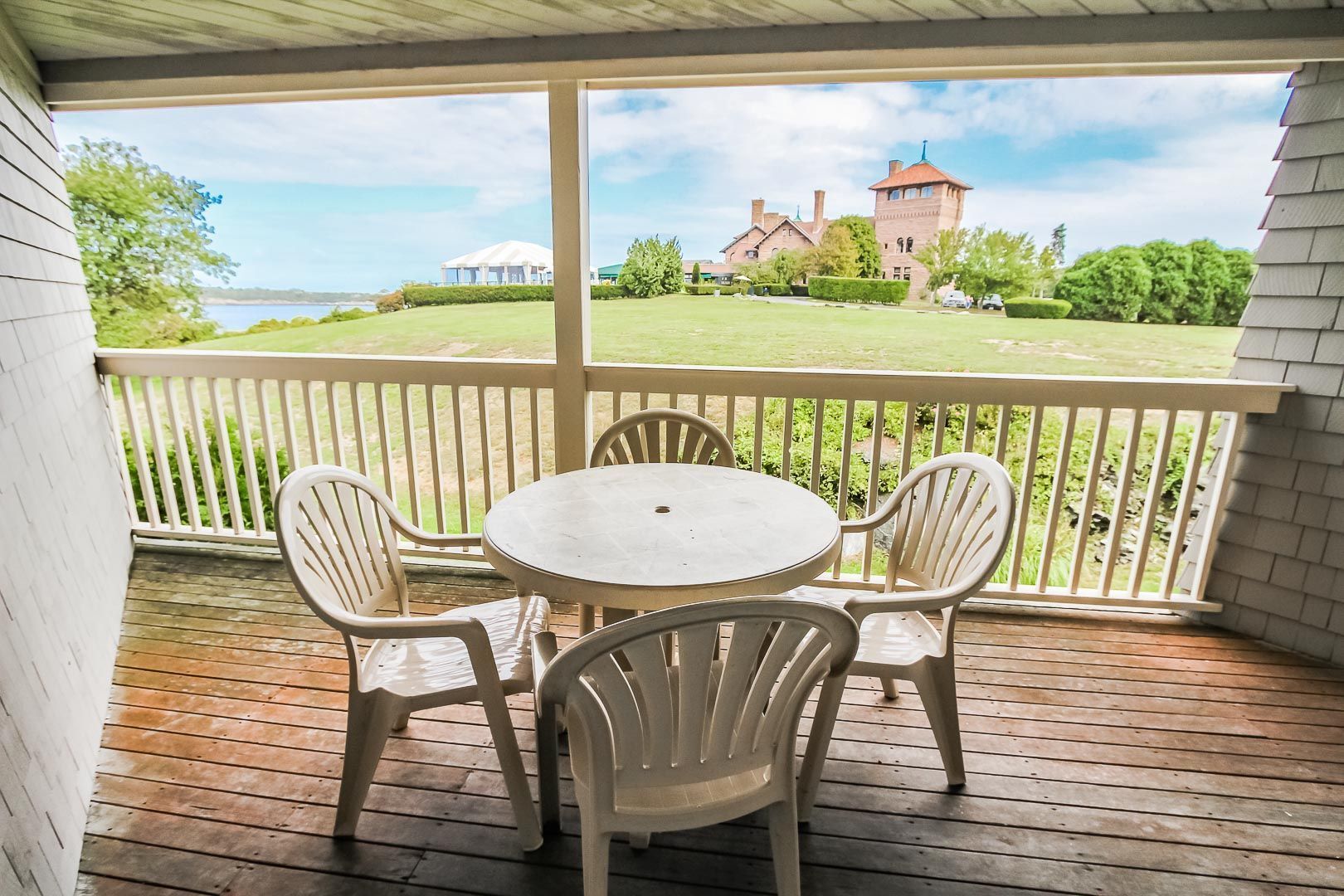 A covered outdoor area with a round table and four chairs, overlooking a well-maintained lawn and a distant building with a tower.