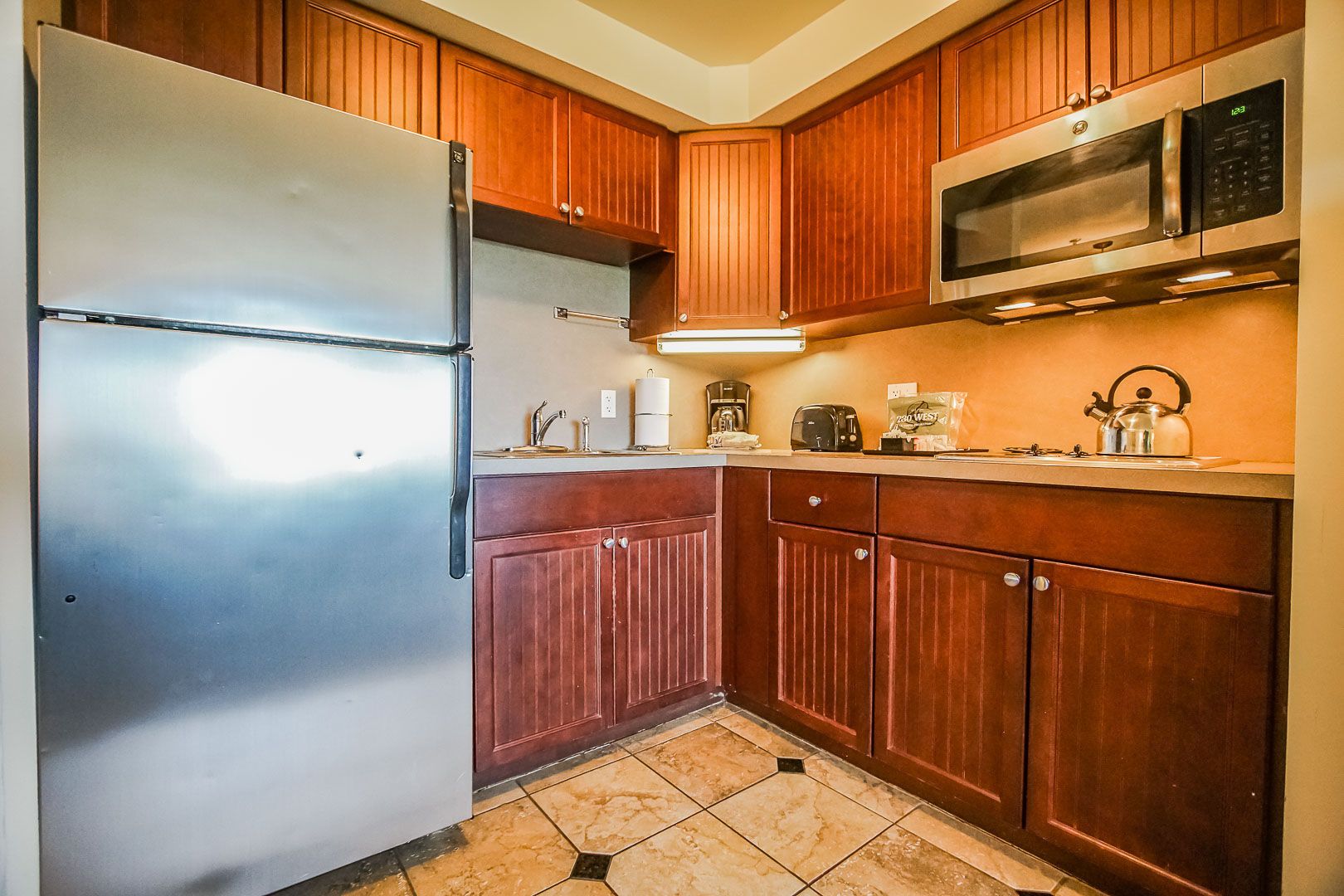 A kitchen with wooden cabinets, a stainless steel refrigerator, a microwave, a kettle, and a coffee maker.