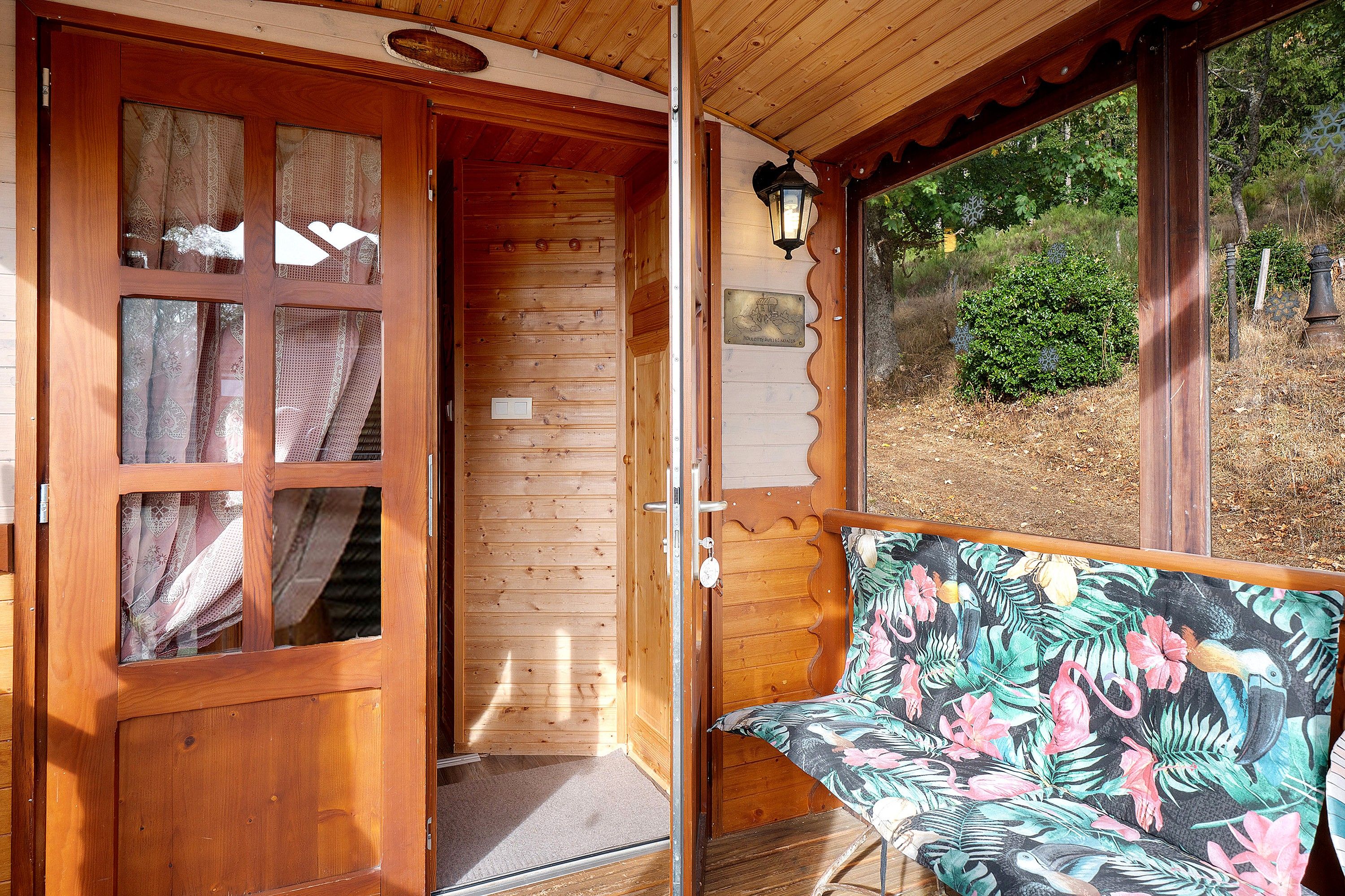 A wooden cabin with a door leading to a room, a bench with a tropical print cushion, and a window with a view of greenery.