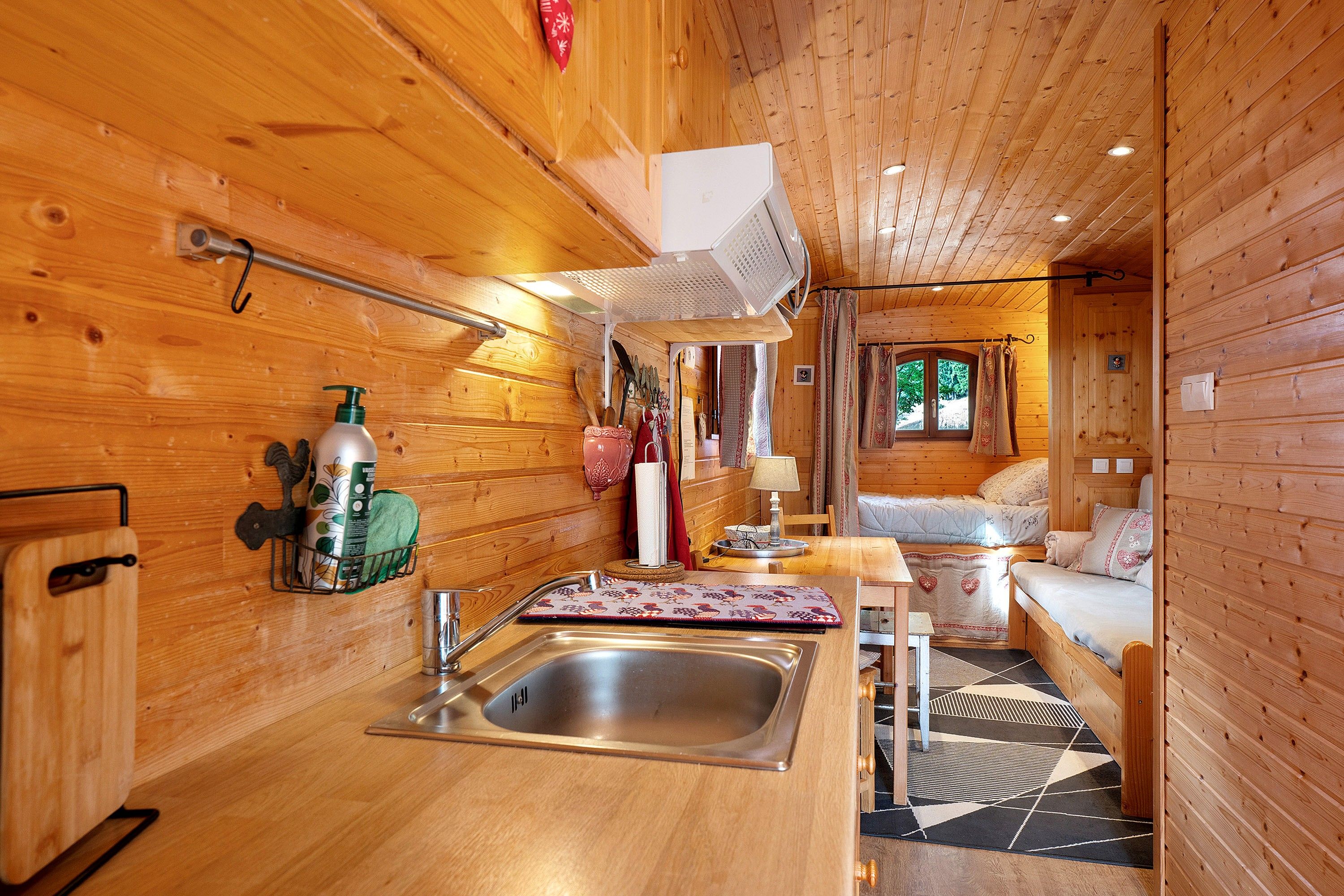 A cozy cabin interior with a wooden interior, a kitchen area with a sink and a hanging rack, and a seating area with a sofa and a window.