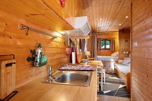 Een knus blokhutinterieur met houten inrichting, een keuken met een spoelbak en een hangrek, en een zithoek met een bank en een raam.