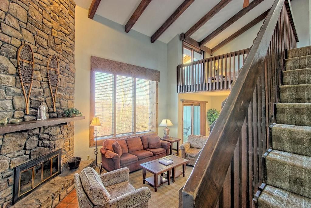 A cozy living room with a stone fireplace, a wooden staircase, and a balcony.