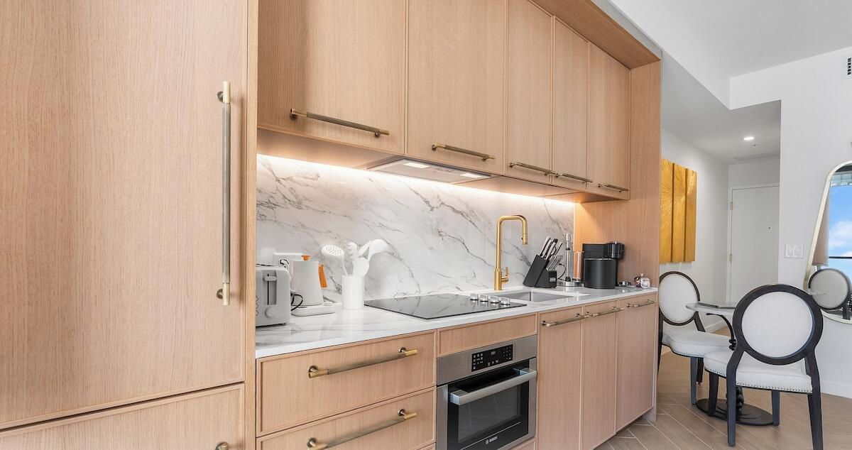 A modern kitchen with wooden cabinets, a marble countertop, a built-in oven, and a dining area with chairs.