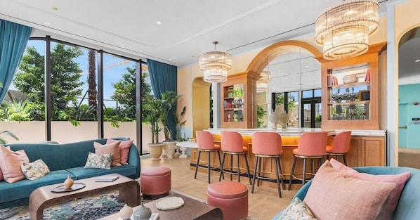 A modern living room with a sofa, coffee table, and bar stools. There is a large window with a view of trees. - Studio w/ Pool & Bar in NoMad Residences Wynwood (Miami)