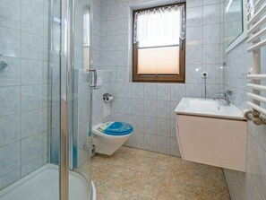 Ein Badezimmer mit Duschkabine aus Glas, Toilette mit blauem Sitz, Waschbecken mit Spiegel und Fenster mit Jalousien.