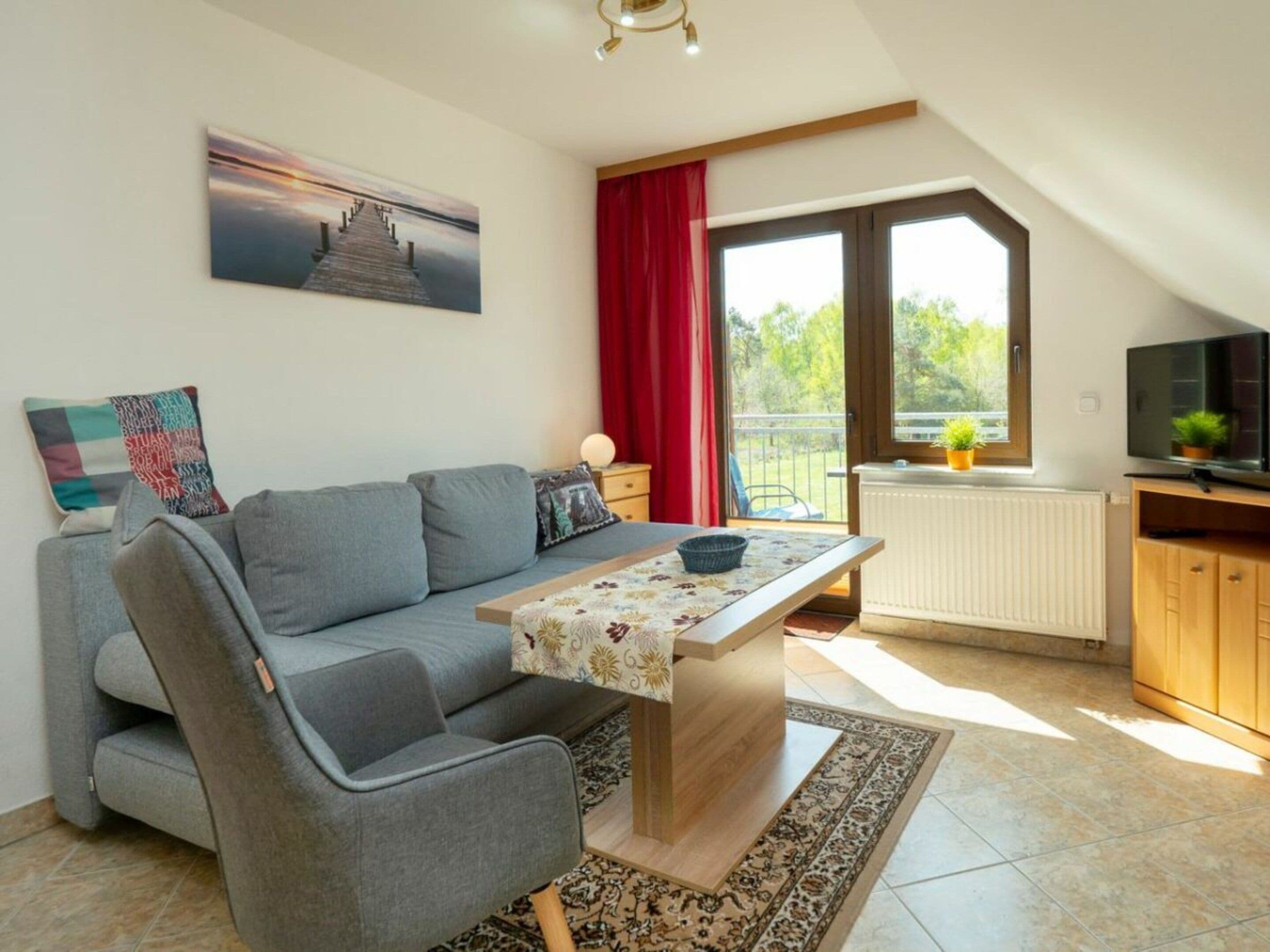 A modern living room with a sofa, a table, a TV, and a large window with a view of greenery.