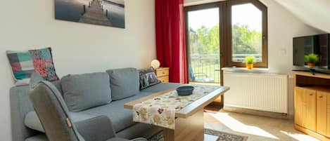 A modern living room with a sofa, a table, a TV, and a large window with a view of greenery.