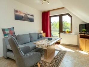 Ein modernes Wohnzimmer mit einer Couch, einem Tisch, einem Fernseher und einem großen Fenster mit Blick auf Grünflächen.
