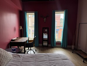 Une pièce comprenant un lit, un bureau, une chaise et deux fenêtres avec des rideaux.