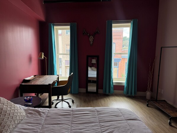 A room with a bed, a desk, a chair, and two windows with curtains.